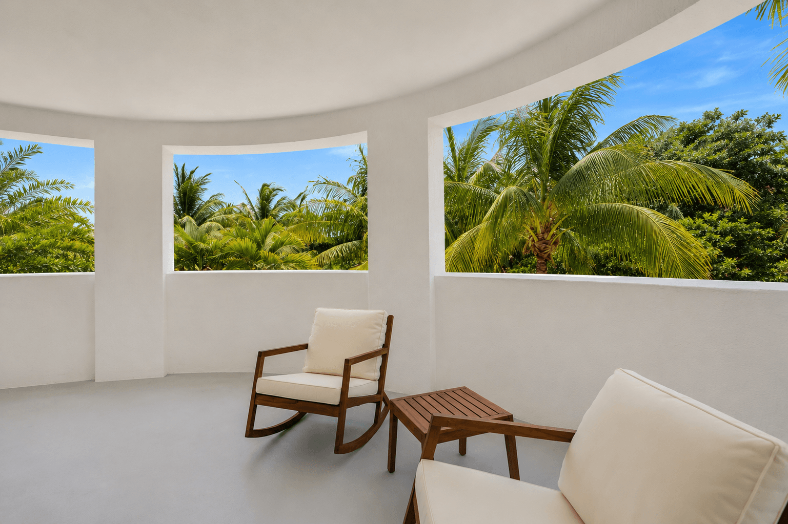 A white chair and table are in a room with windows overlooking a tropical landscape.
